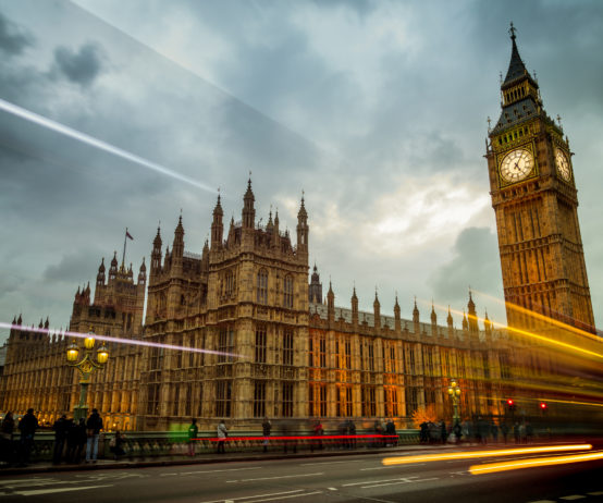 Big Ben and the Houses of Parliament in London, England. The building is lit up and set against a dramatic sky with cars and busses zooming by on Waterloo Bridge.