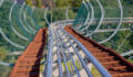 Curved metal roller coaster track extends downward amidst trees on a sunny day.
