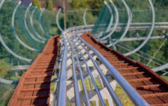 Curved metal roller coaster track extends downward amidst trees on a sunny day.