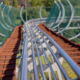 Curved metal roller coaster track extends downward amidst trees on a sunny day.