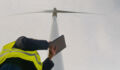 Engineer wearing safety vest holding digital tablet, collecting data and monitoring operation of wind turbine for green energy production against cloudy sky