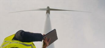 Engineer wearing safety vest holding digital tablet, collecting data and monitoring operation of wind turbine for green energy production against cloudy sky