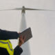 Engineer wearing safety vest holding digital tablet, collecting data and monitoring operation of wind turbine for green energy production against cloudy sky