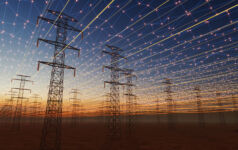 A futuristic visualization of high-voltage transmission towers beneath a digital energy grid. Glowing network lines and light points span across the sky, symbolizing smart grid technology, connectivity, and the modernization of electrical infrastructure at sunset.