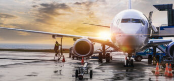 An aeroplane at an airport being refuelled and loaded as the sun sets or rises over the horizon